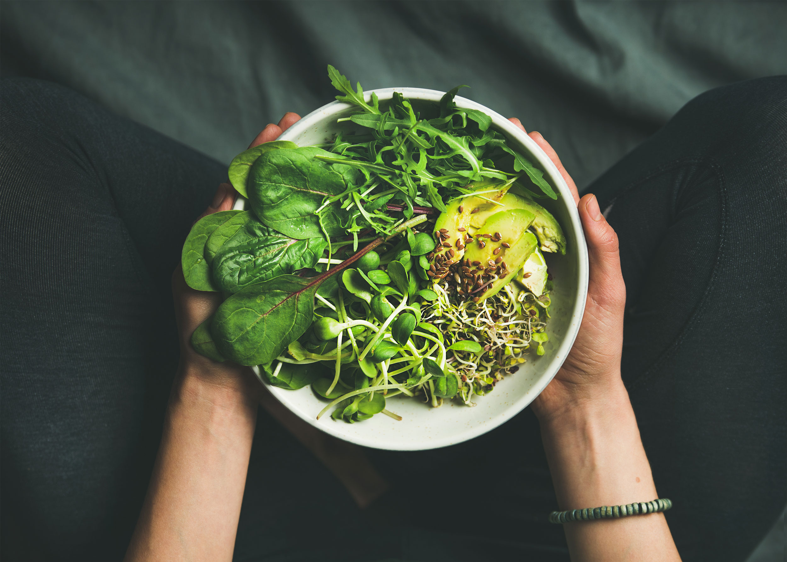 A person is holding a bowl of fresh green salad, avocado and sprouts on their lap whilst sitting on a dark piece of fabric.