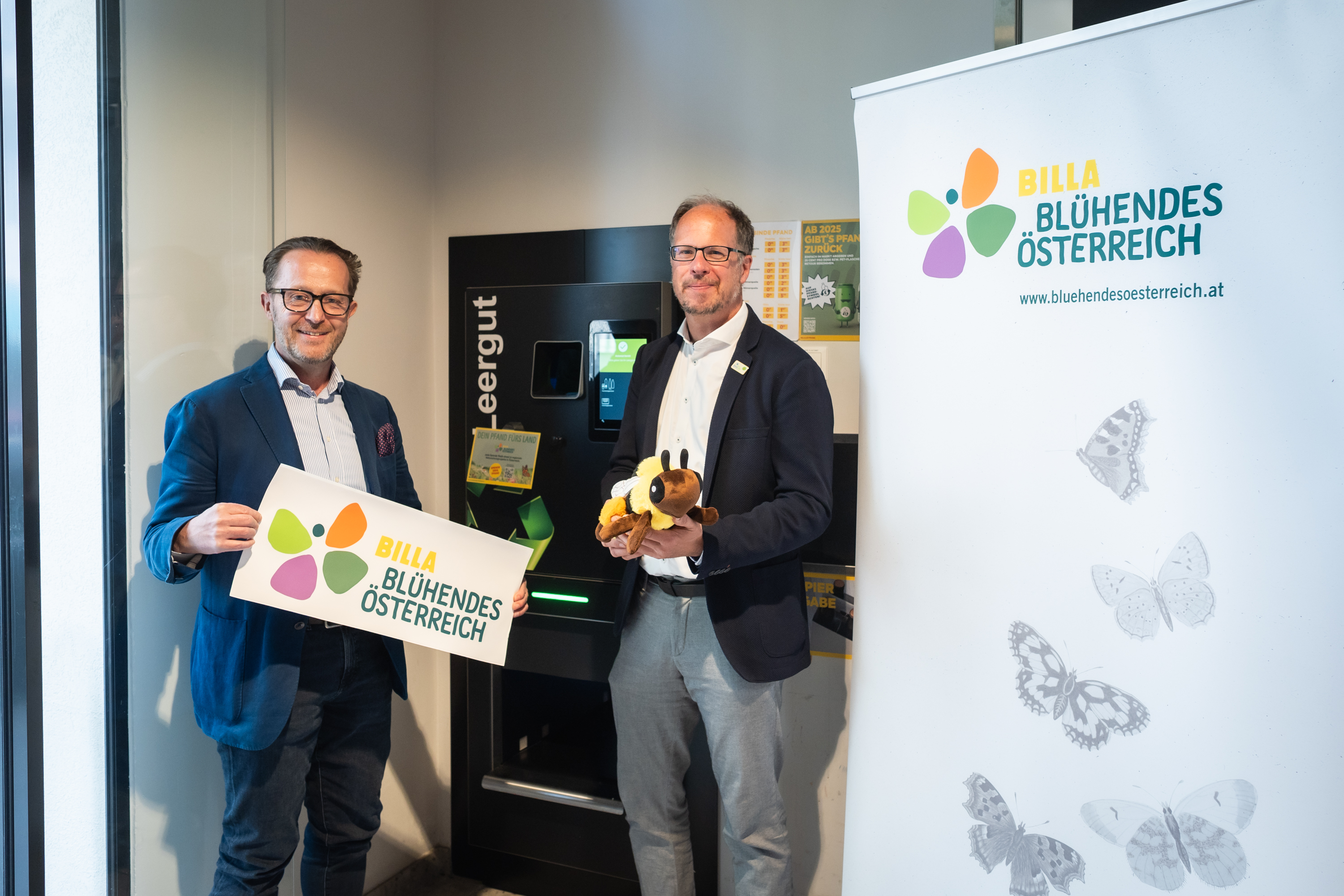 Two people in suits are standing in front of a bottle return machine, holding a soft toy and a sign bearing the BILLA &lsquo;Bl&uuml;hendes &Ouml;sterreich&rsquo; logo and lettering.