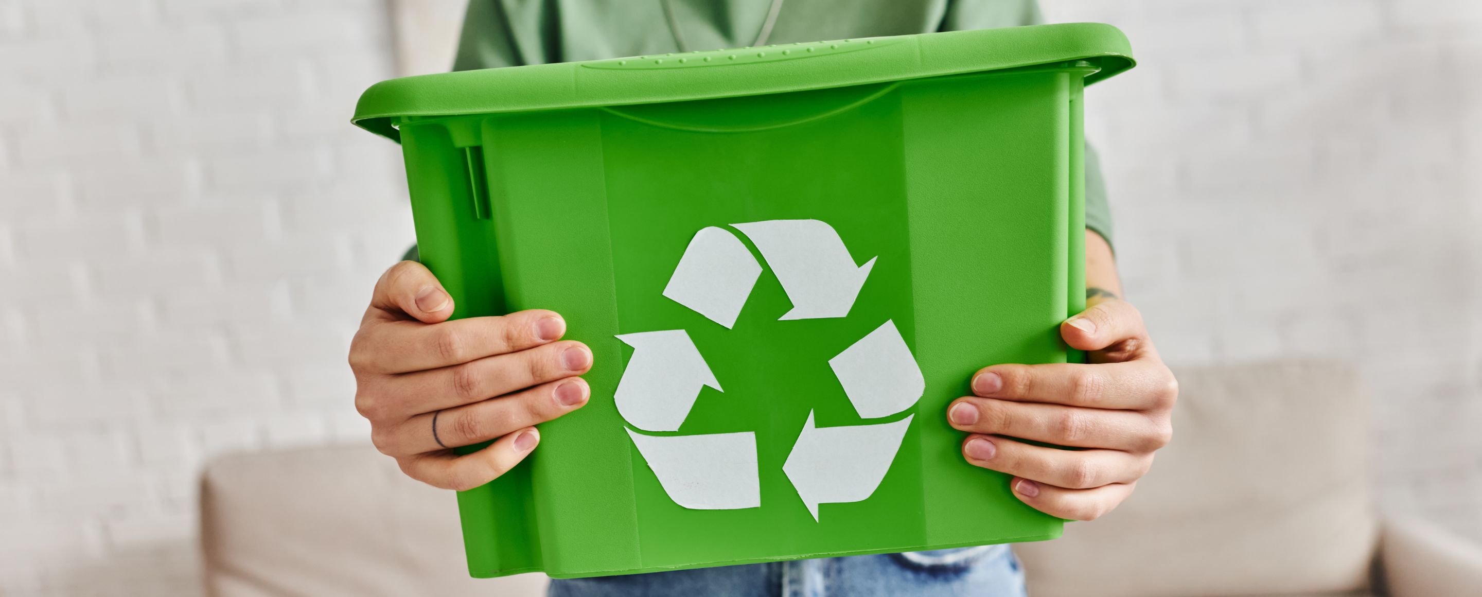A person wearing a green top and jeans is holding a green recycling bin with the white recycling symbol in front of a light-coloured sofa in a room with a white brick wall.