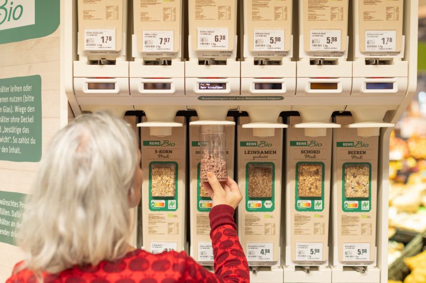 A person with grey hair and a red jumper is filling a container with organic grains from a dispenser in a supermarket.