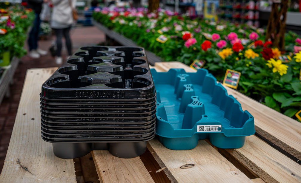 On a wooden table in a garden centre, stacks of black and blue reusable planters stand alongside colourful flowers and plants.