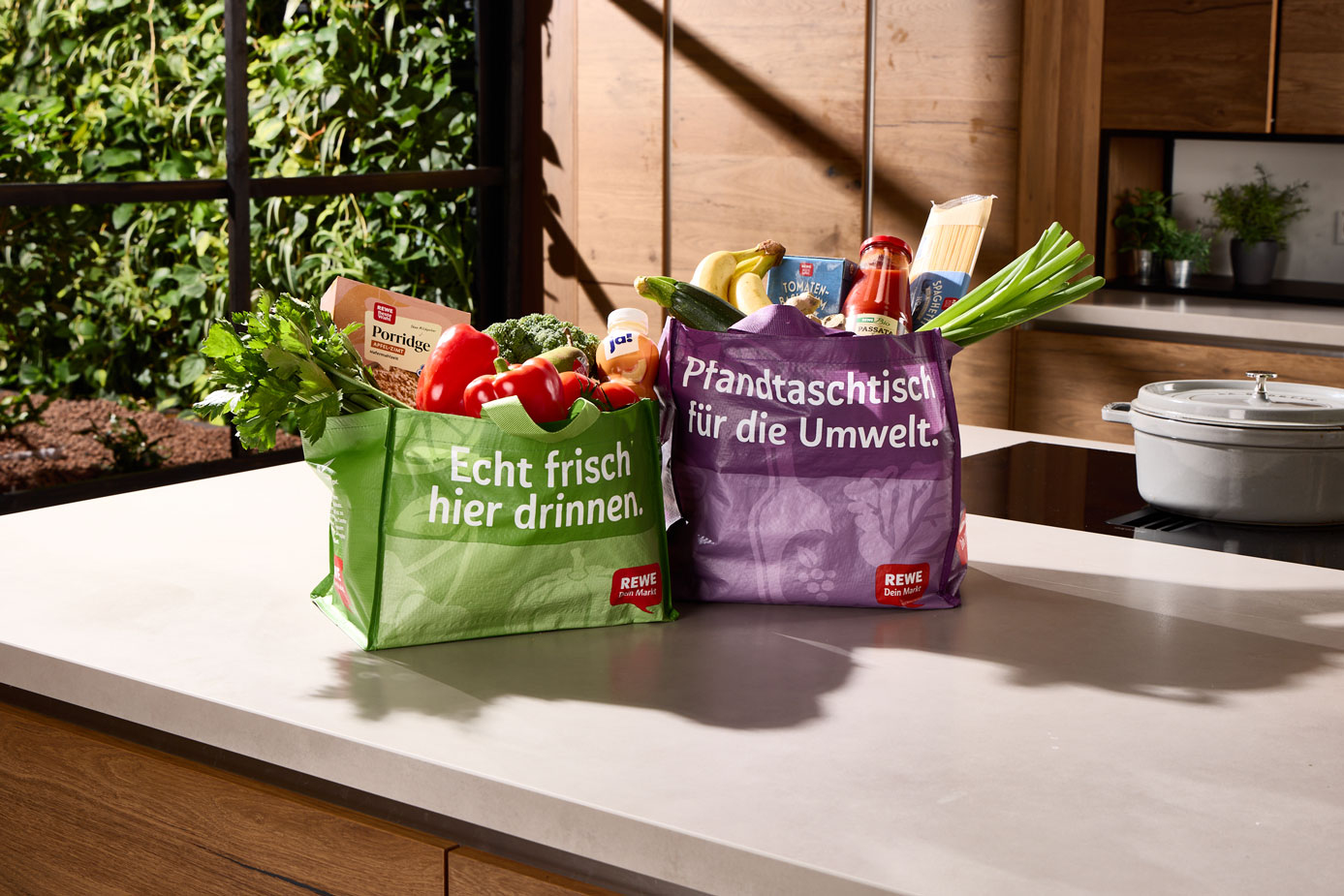 Two full reusable shopping bags filled with fresh vegetables and groceries are sitting on a kitchen worktop in front of a window overlooking the countryside.