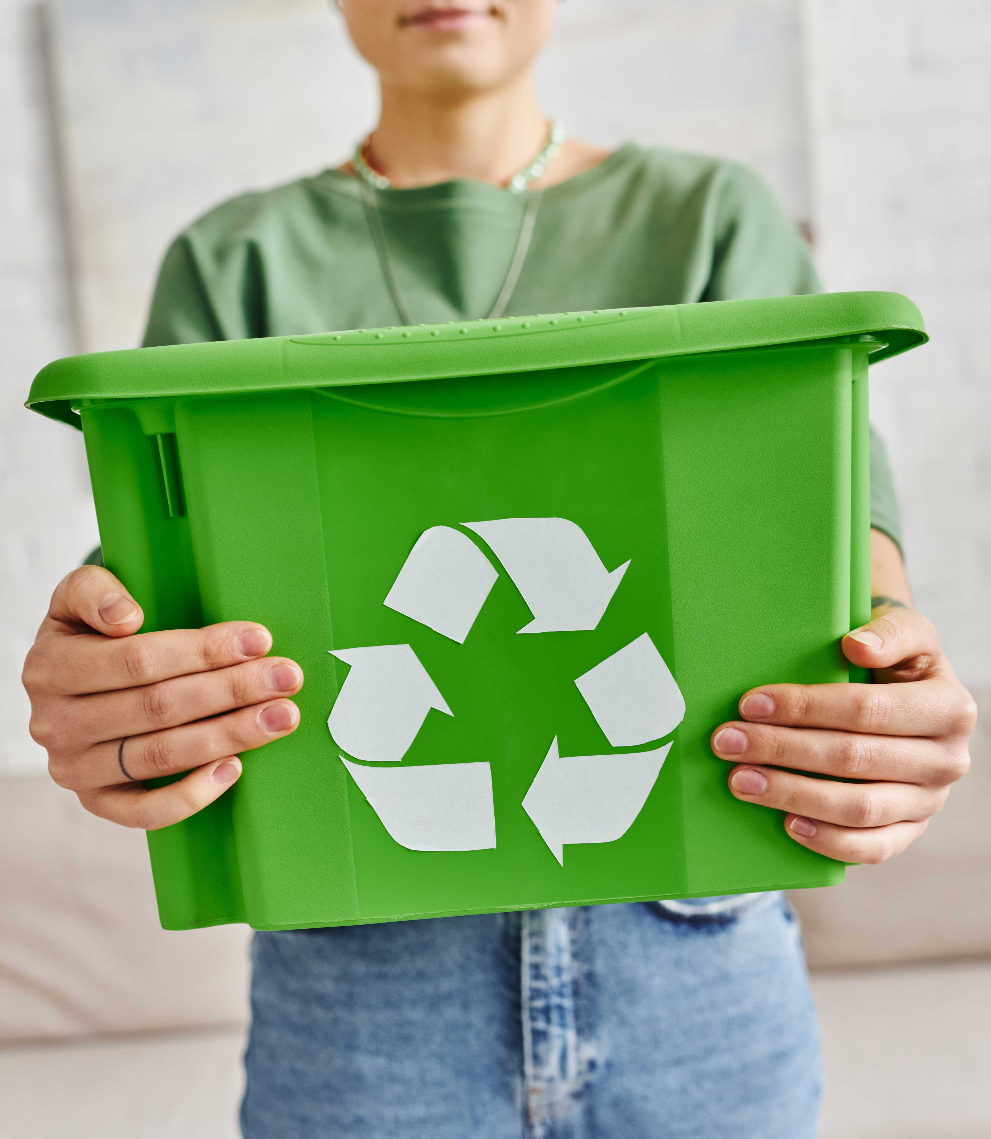 A person wearing a green top and jeans is holding a green recycling bin with the white recycling symbol in front of a light-coloured sofa in a room with a white brick wall.