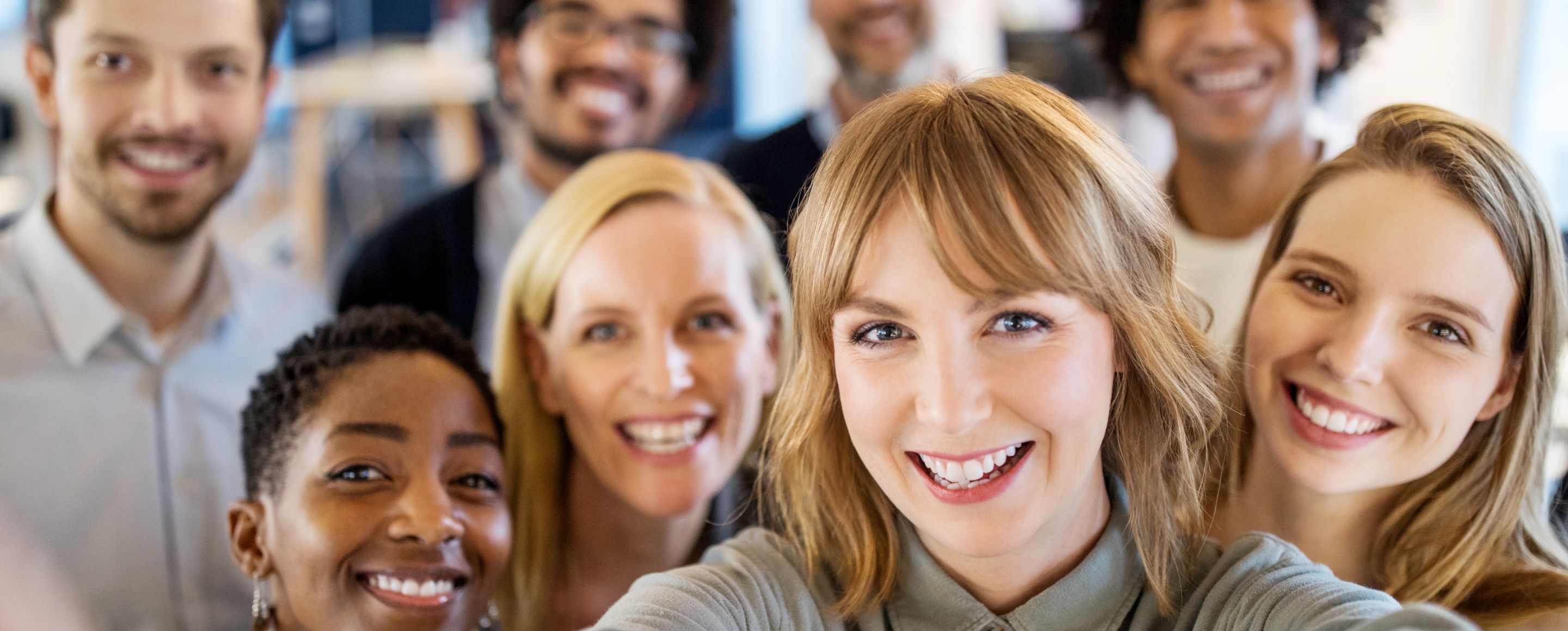 A group of people are standing close together and smiling at the camera, whilst someone in the foreground is taking a selfie.