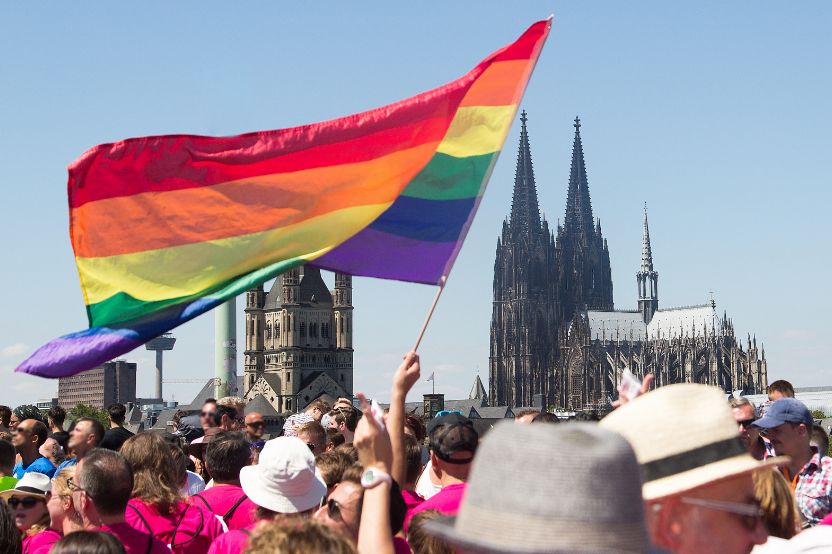 A crowd of people in pink T-shirts are gathered outdoors, whilst one person is waving a large rainbow flag in front of a Gothic cathedral.