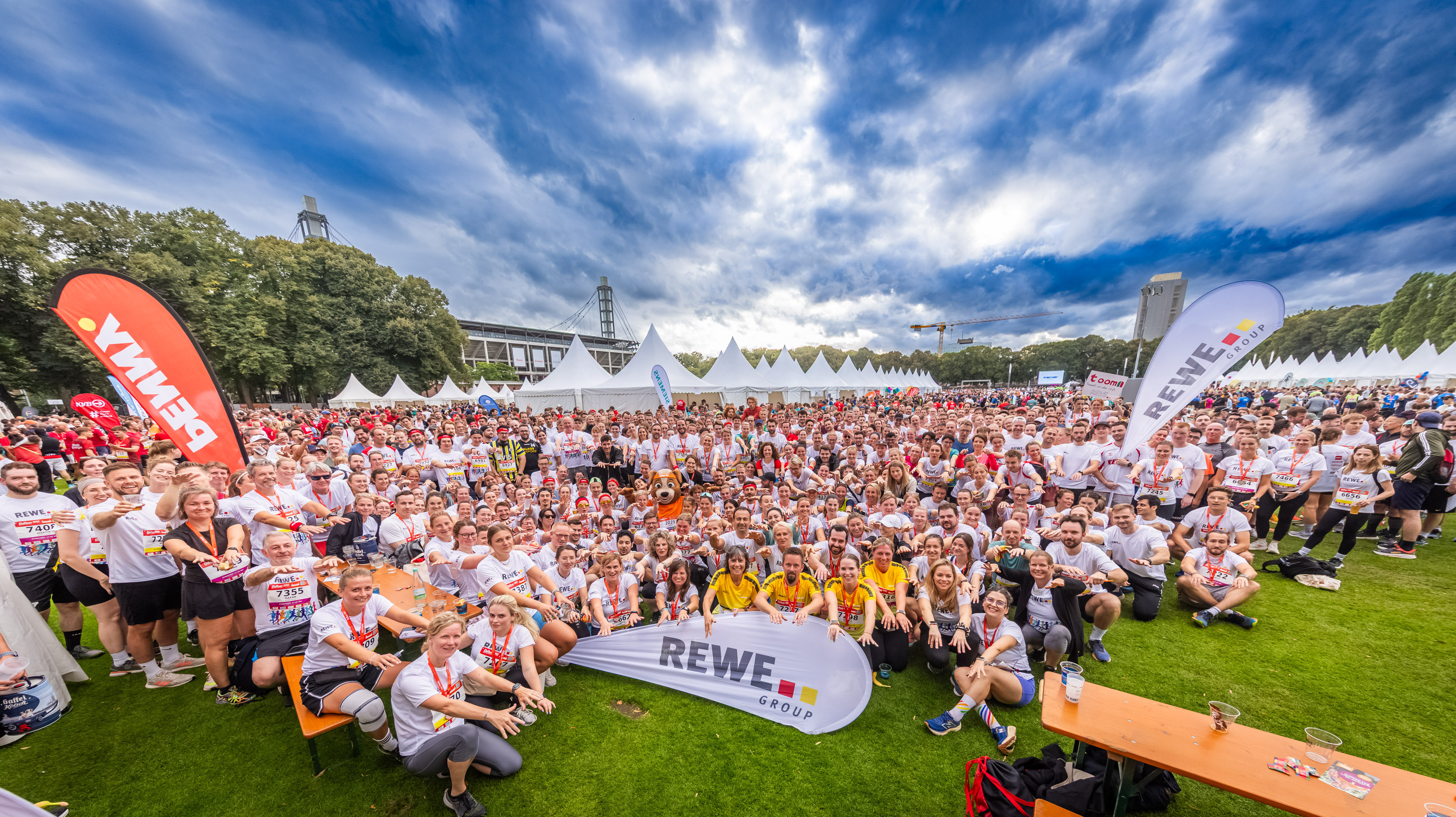 A large group of people in sportswear are gathered on a lawn in front of white tents under a cloudy sky; some are holding banners bearing the word &lsquo;REWE&rsquo;.