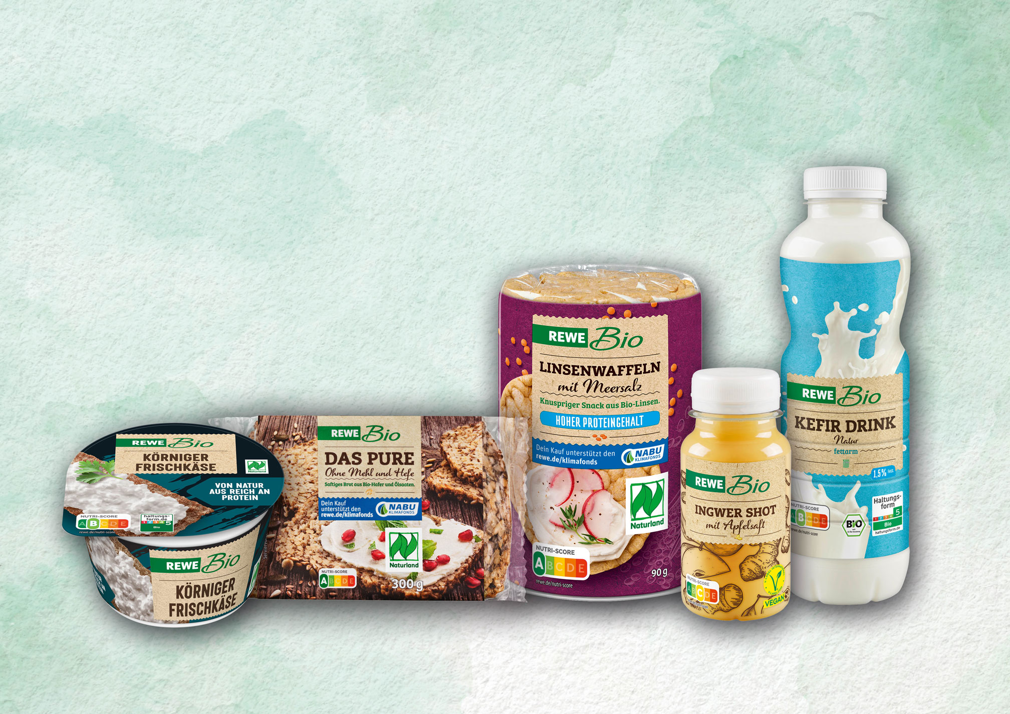 Several organic food products from the REWE Bio range are arranged side by side against a light-coloured background, including cottage cheese, bread, lentil waffles, a ginger shot and a kefir drink.