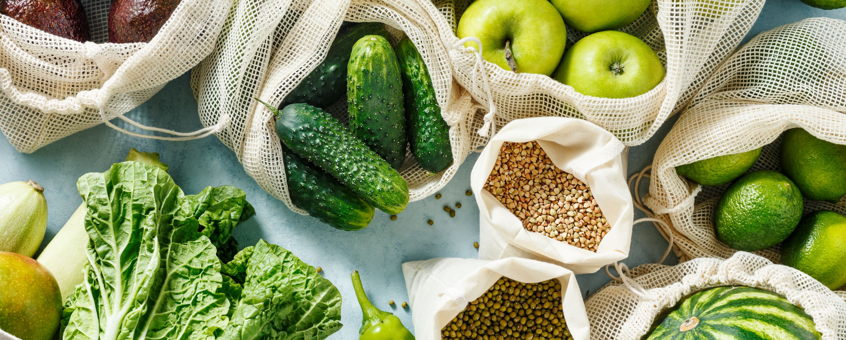 Various fresh foods, such as lettuce, gherkins, apples, green chillies and pulses, are arranged on a light-coloured surface in reusable mesh bags and cloth bags.