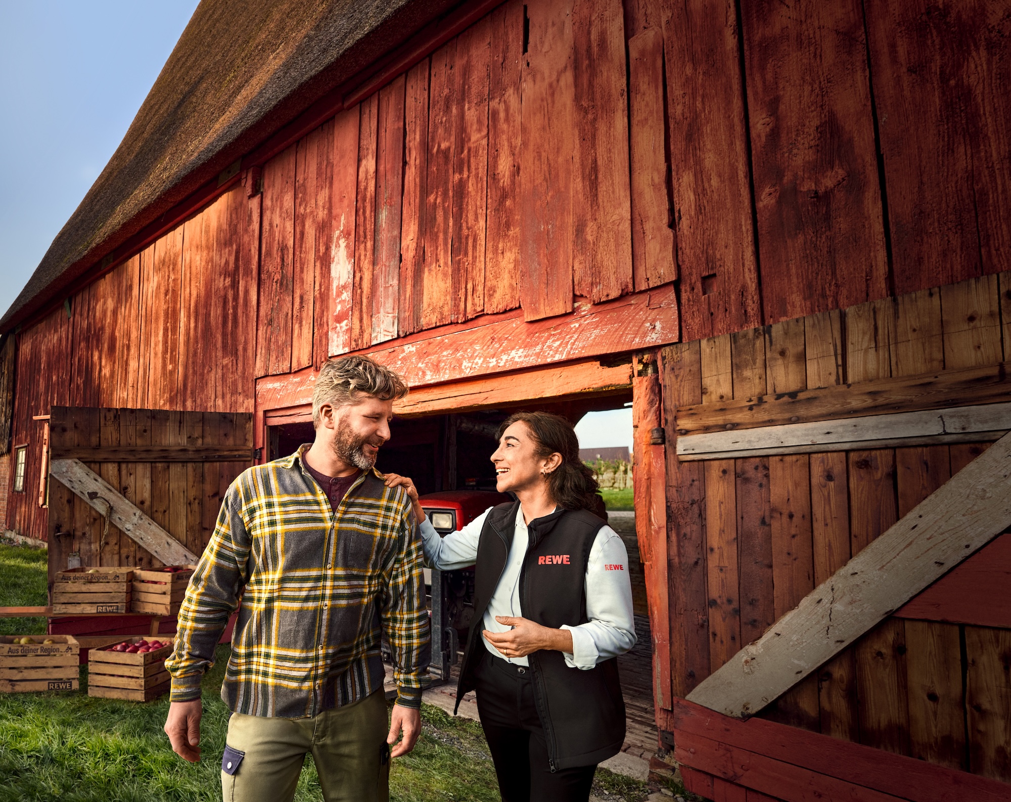 Two people are standing in front of a large red wooden building with its doors open; one is wearing a checked shirt and the other a black waistcoat, and they appear to be having a friendly chat.