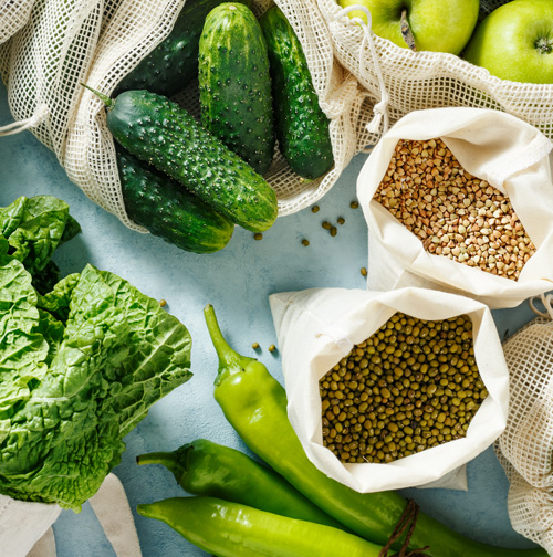 Various fresh foods, such as lettuce, gherkins, apples, green chillies and pulses, are arranged on a light-coloured surface in reusable mesh bags and cloth bags.