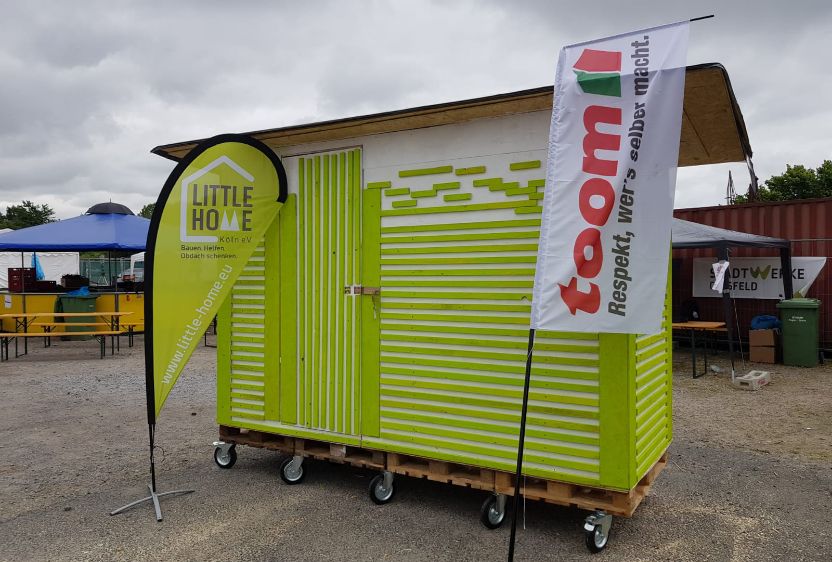 A small mobile site hut with green stripes is parked in a car park next to two flags bearing the words &lsquo;Little Home&rsquo; and &lsquo;toom&rsquo;.