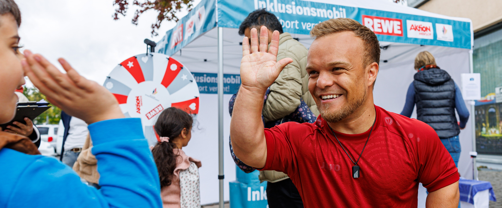 Several people are standing in front of a stall with a wheel of fortune, and one person in a red shirt is raising their hand in greeting