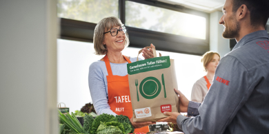 A person wearing an orange apron with the Tafel logo is handing a paper bag bearing the words &lsquo;Filling plates together&rsquo; to another person in a brightly lit room, with vegetables in the foreground.