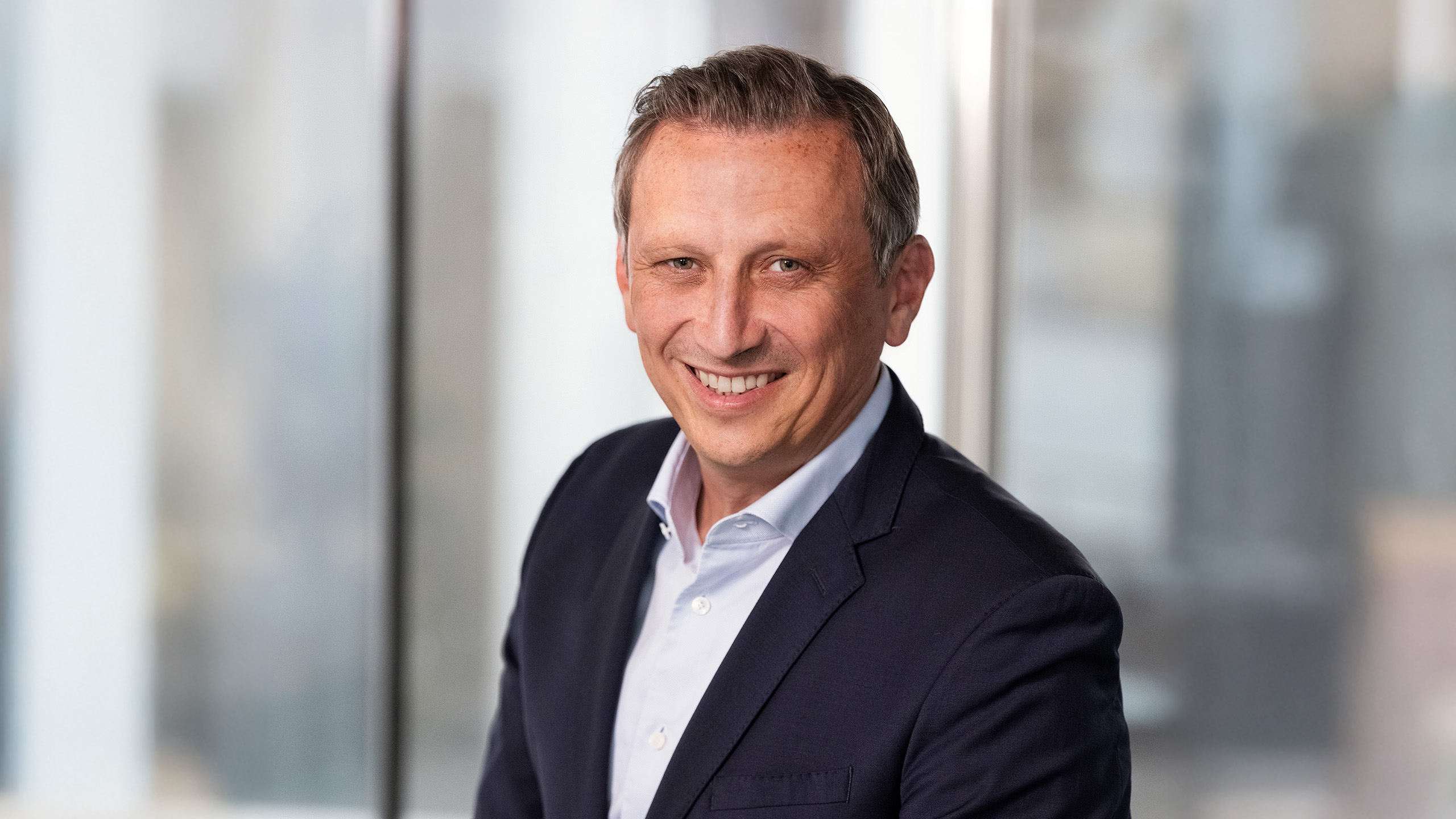Lionel Souque, CEO of the REWE Group, is wearing a white shirt and a dark suit and is standing in front of a window with a blurred background.