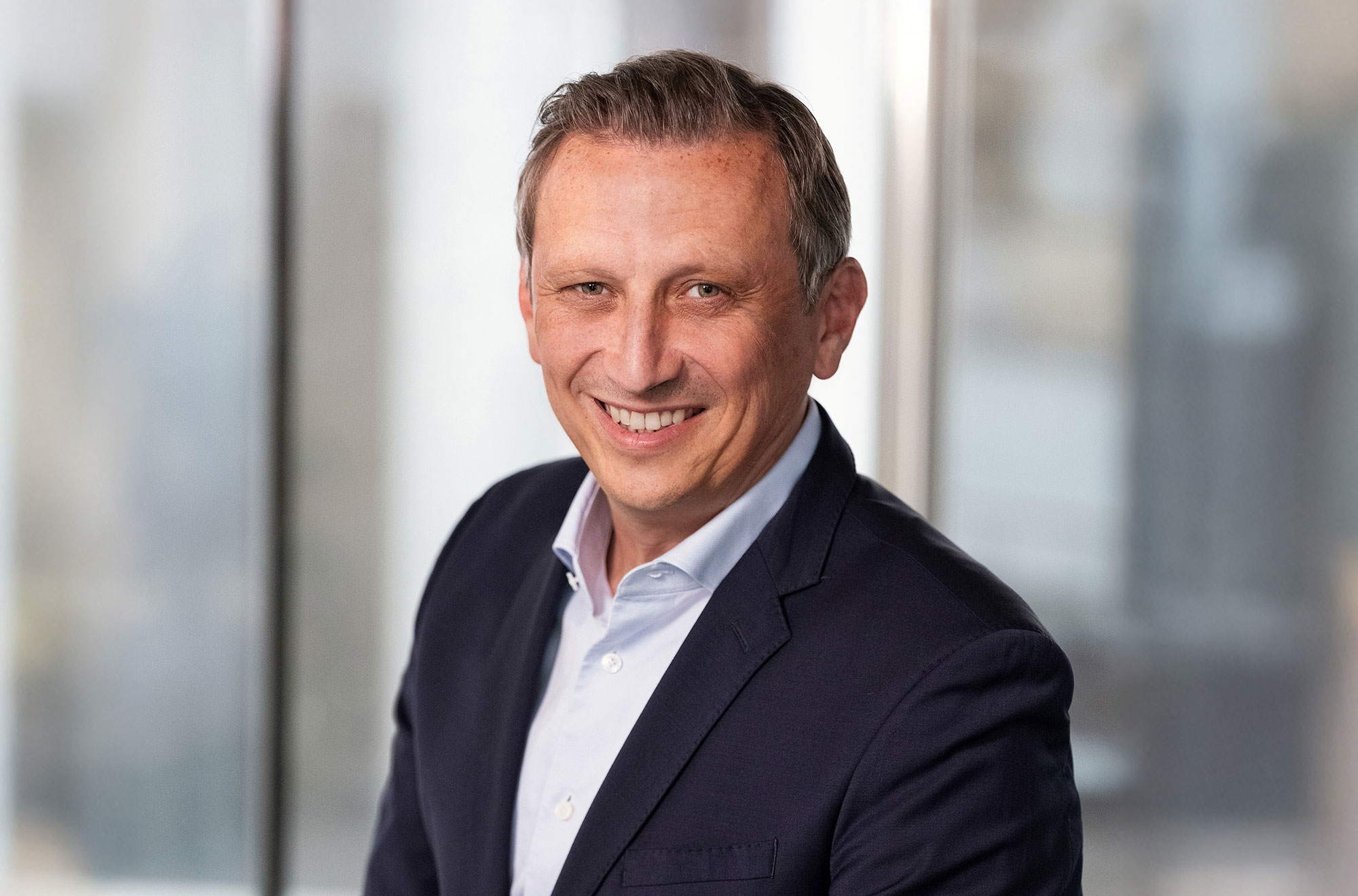 Lionel Souque, CEO of the REWE Group, is wearing a white shirt and a dark suit and is standing in front of a window with a blurred background.