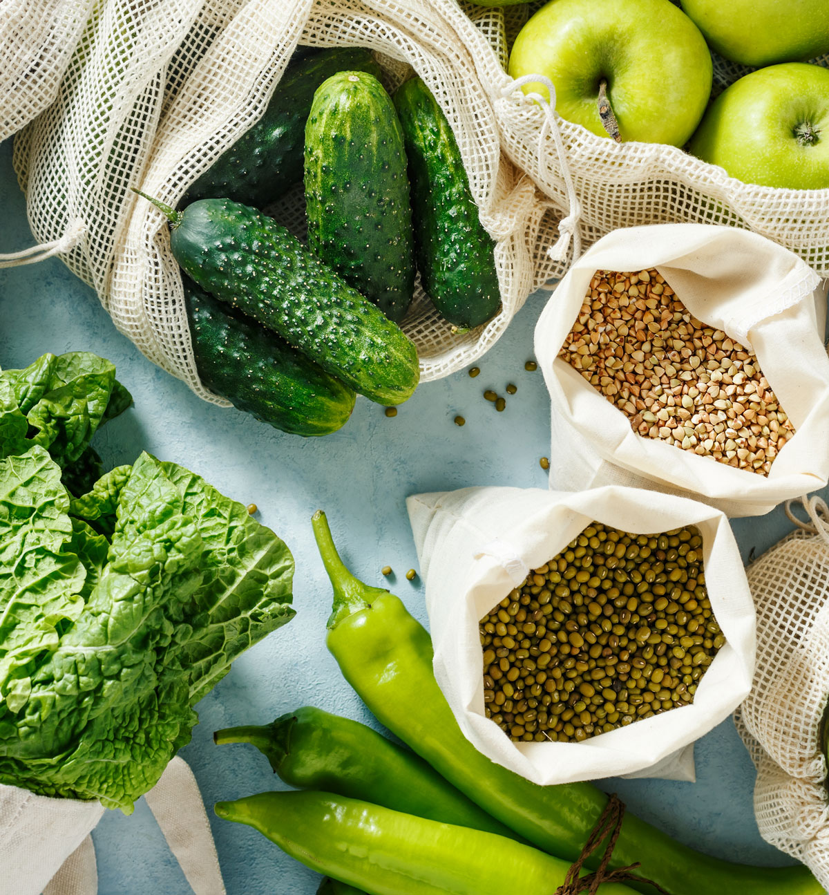 Verschiedene frische Lebensmittel wie Salat, Gurken, Äpfel, grüne Chilischoten und Hülsenfrüchte sind in wiederverwendbaren Netztaschen und Stoffbeuteln auf einer hellen Oberfläche arrangiert.