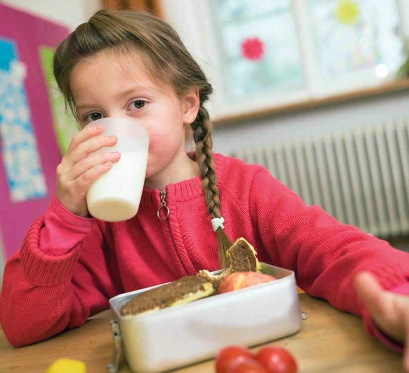 Ein Kind mit geflochtenem Haar und rotem Pullover sitzt an einem Tisch und trinkt aus einem Becher, während es vor sich eine Brotdose mit Essen hat.