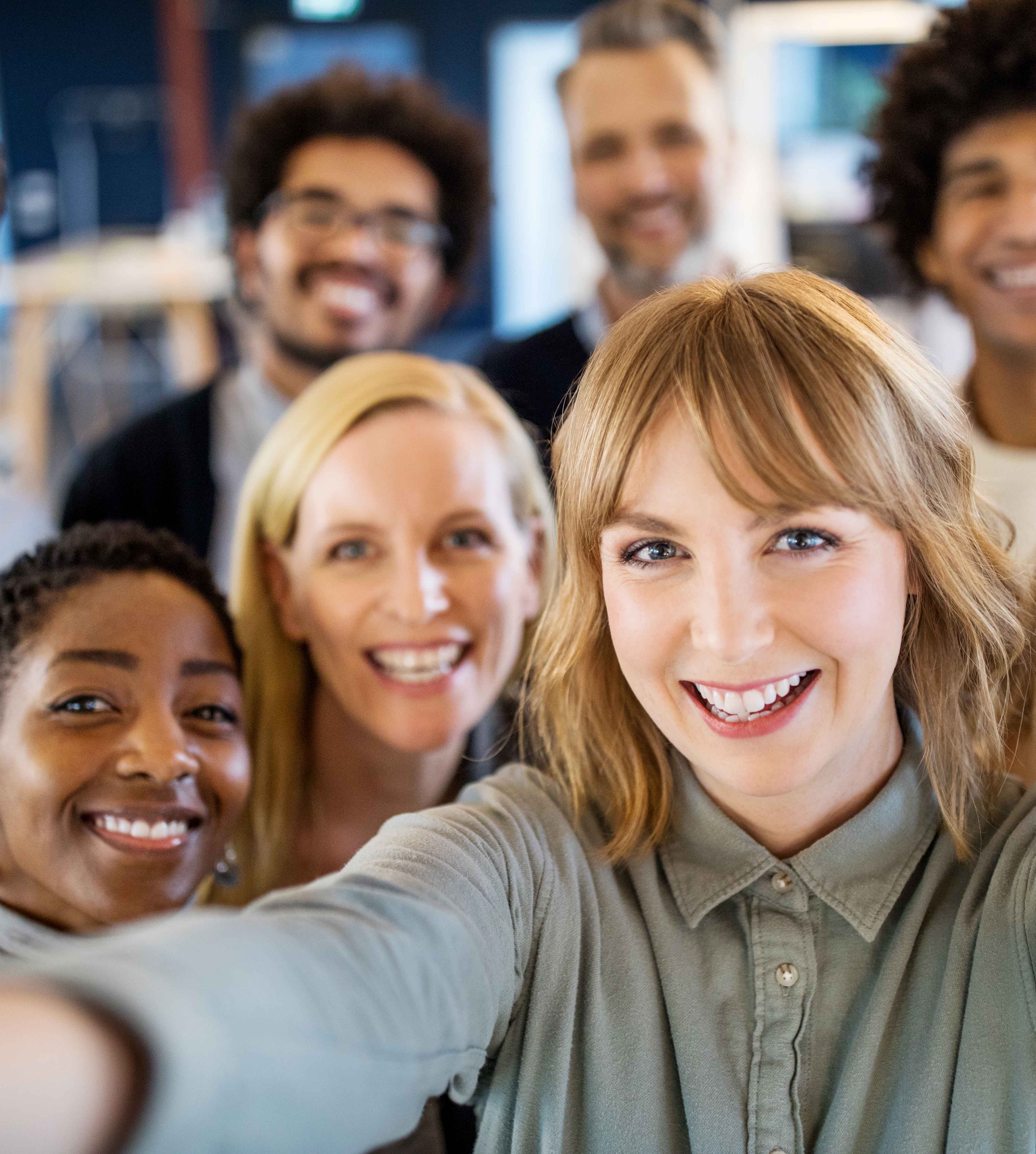 Eine Gruppe von Menschen steht eng zusammen und lächelt in die Kamera, während eine Person im Vordergrund einen Selfie macht.