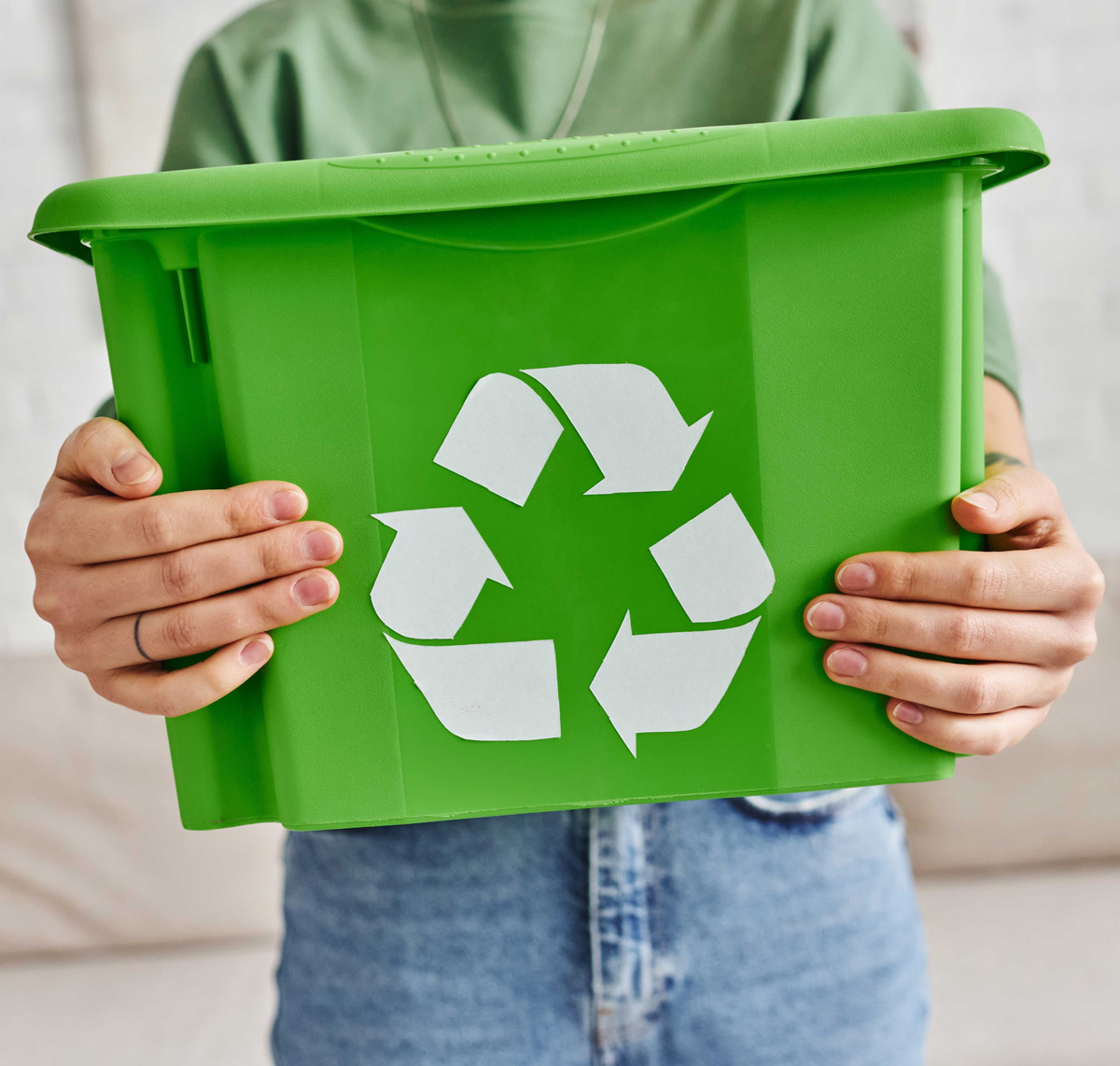 Eine Person in grünem Oberteil und Jeans hält eine grüne Recyclingbox mit dem weißen Recycling-Symbol vor einem hellen Sofa in einem Raum mit weißer Ziegelwand.