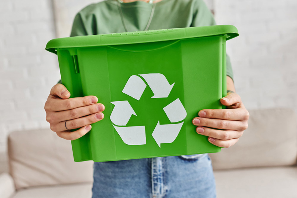 A person wearing a green top and jeans is holding a green recycling bin with the white recycling symbol in front of a light-colored sofa in a room with a white brick wall.
