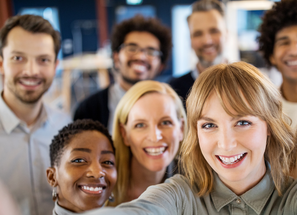 A group of people is standing close together and smiling at the camera while one person in the foreground is taking a selfie.