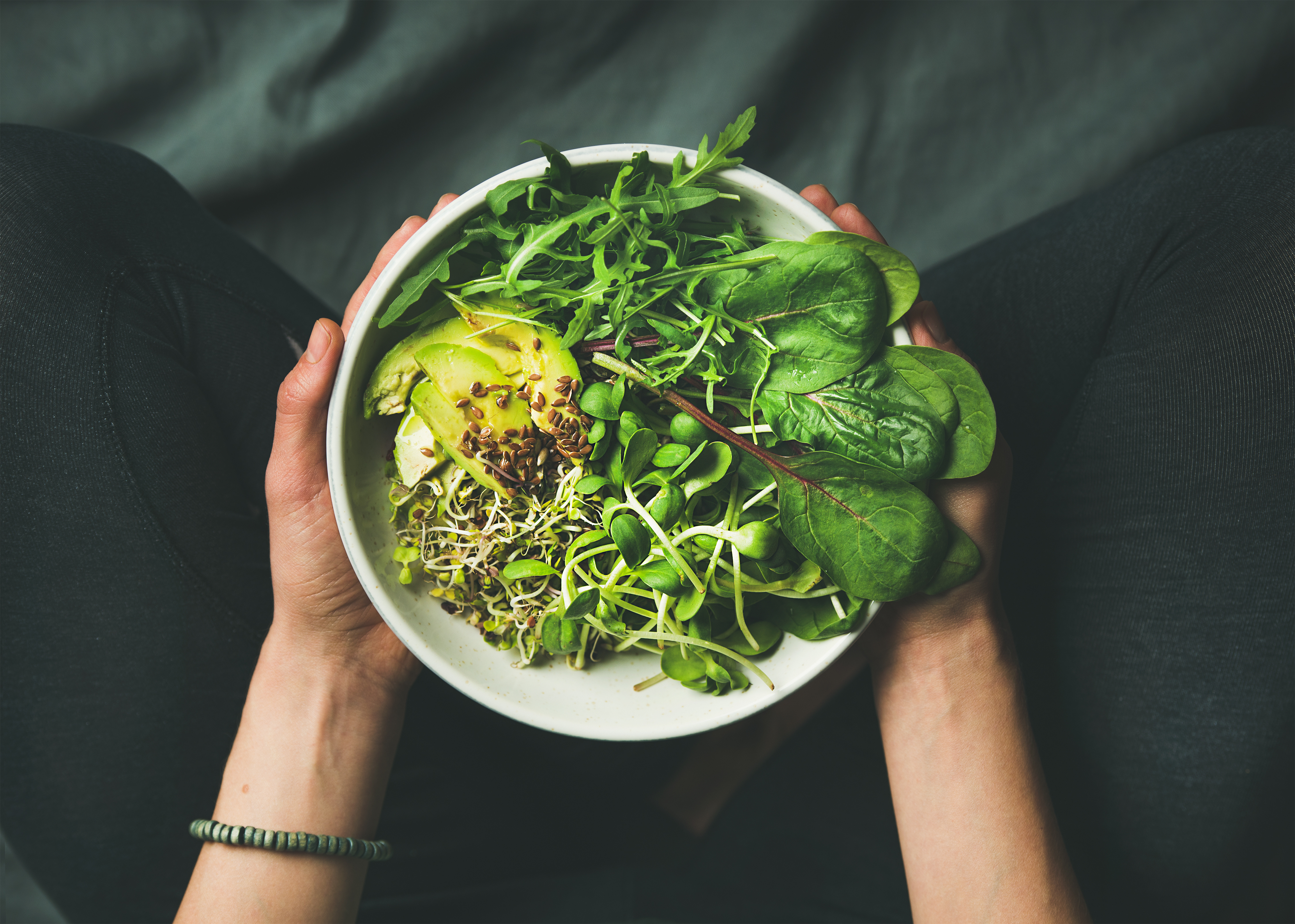 A person is holding a bowl of fresh green salad, avocado, and sprouts on their lap while sitting on a dark piece of fabric.