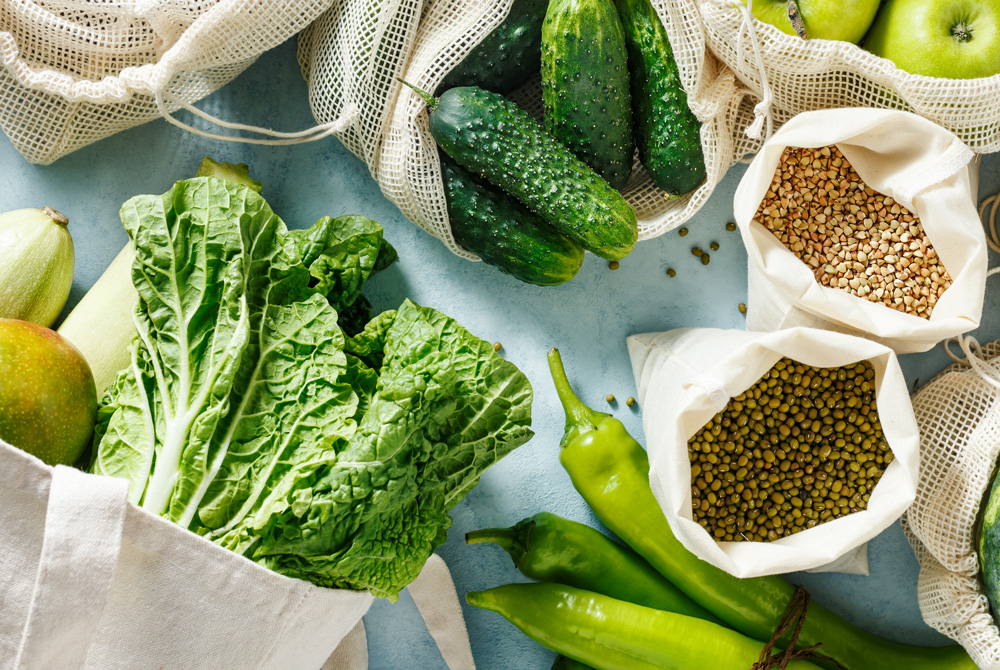 Various fresh foods, such as lettuce, cucumbers, apples, green chili peppers, and legumes, are arranged on a light-colored surface in reusable mesh bags and cloth bags.