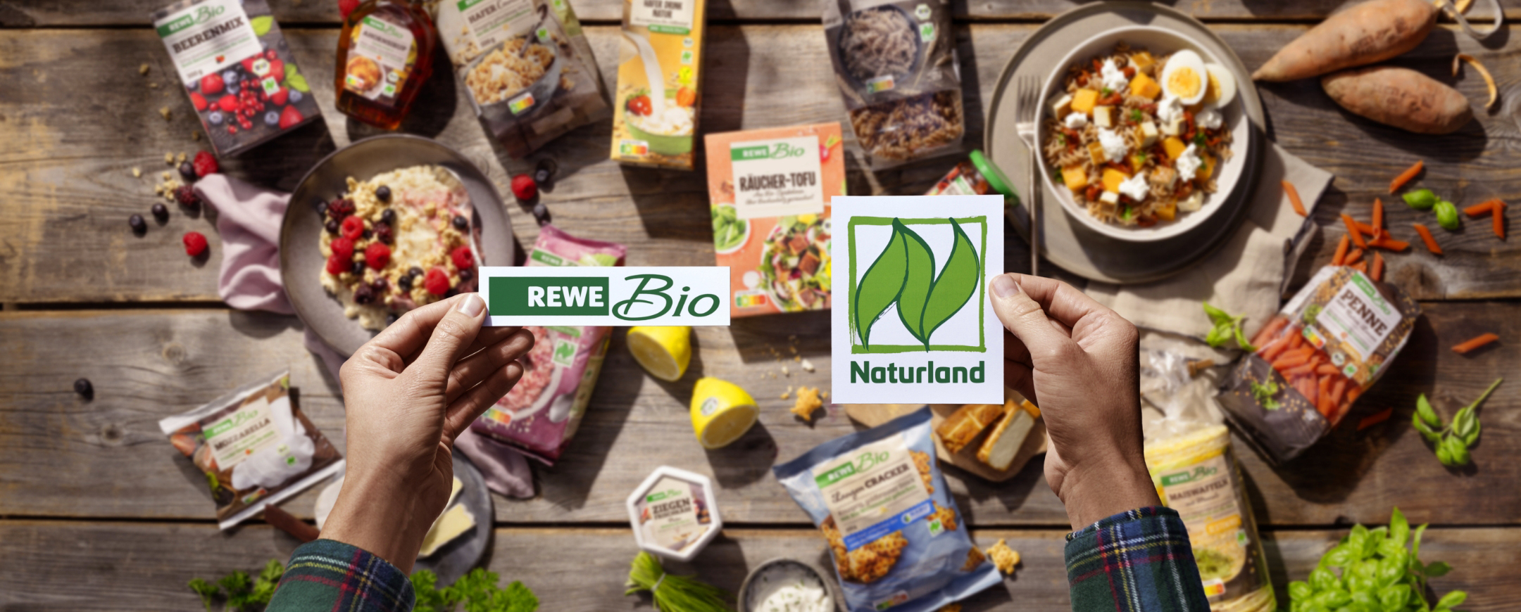 Various organic food products from the REWE Bio range, such as muesli, yoghurt with berries, eggs, herbs, nuts and juices, are arranged attractively on a rustic wooden table.